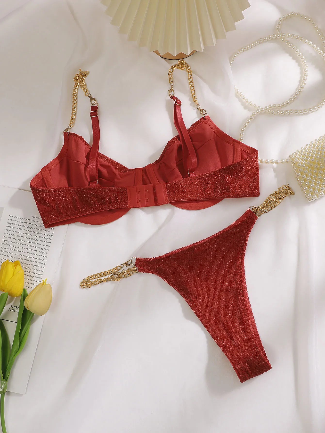 A red satin and textured lace lingerie set with gold chain straps, laid flat on a white fabric background with decorative props including yellow tulips, a pearl necklace, and a folded paper fan.