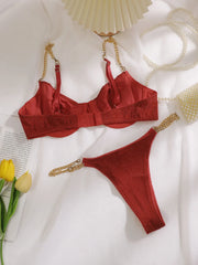 A red satin and textured lace lingerie set with gold chain straps, laid flat on a white fabric background with decorative props including yellow tulips, a pearl necklace, and a folded paper fan.