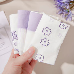 Hand holding a pack of white sanitary pads with purple flower designs on a neutral background.