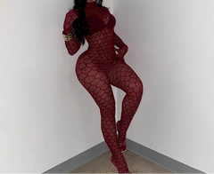 A woman wearing a long-sleeve, high-neck, full-body red fishnet jumpsuit with a textured pattern, posing in a minimalist white room corner.