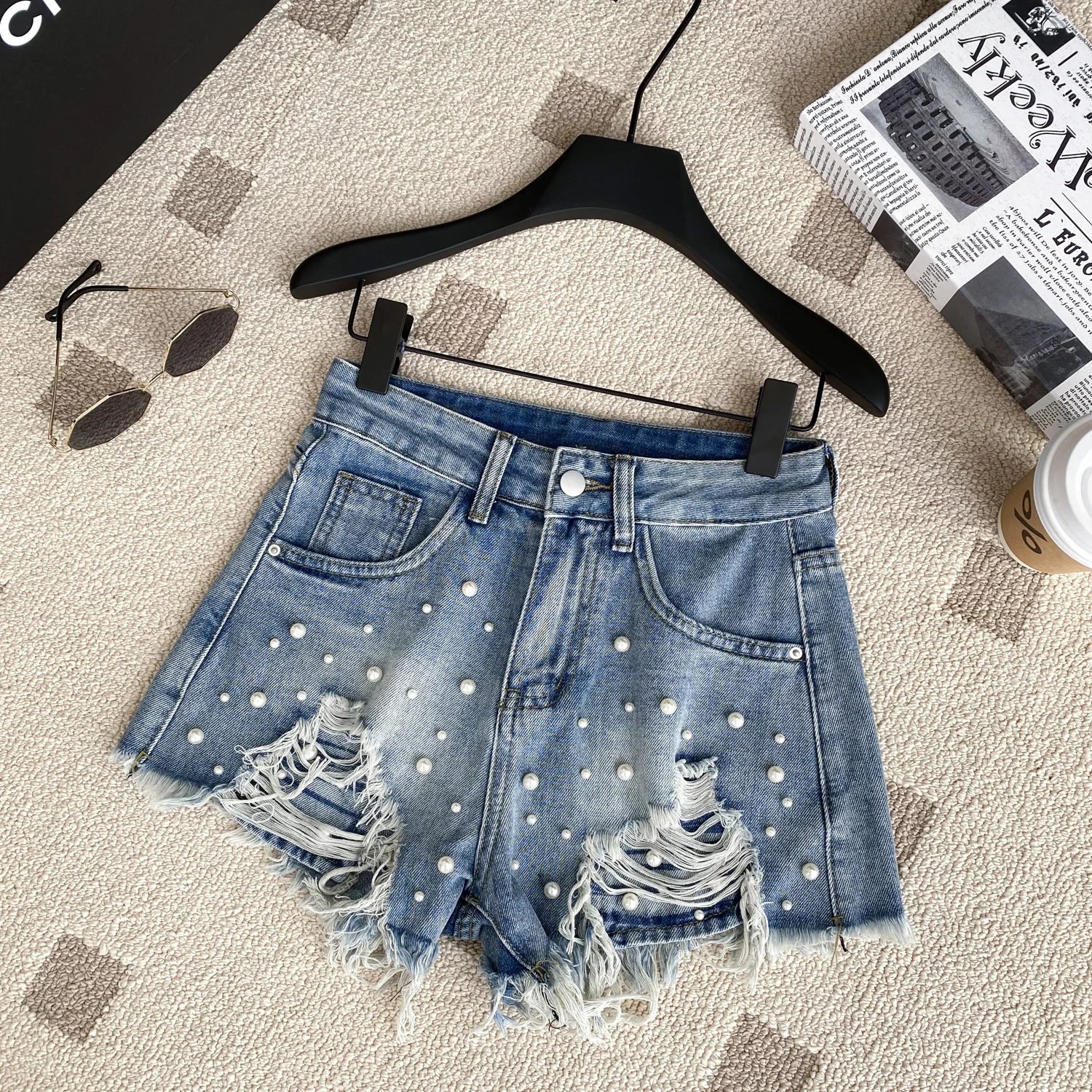 Denim shorts with pearl embellishments on a hanger with sunglasses, newspaper, and cup in the background.