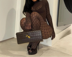 A woman wearing a dark brown patterned long-sleeve bodysuit and matching fishnet tights, posing with a woven black handbag and black platform heels in a minimalist indoor setting.