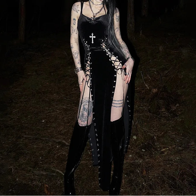 Person in a dark forest wearing a black dress with lace-up front and cross necklace.