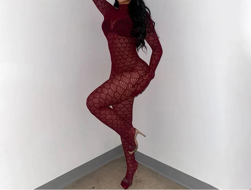 A woman poses in a room corner wearing a long-sleeve, full-body burgundy lace bodysuit with matching gloves and high heels.