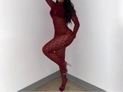 A woman poses in a room corner wearing a long-sleeve, full-body burgundy lace bodysuit with matching gloves and high heels.