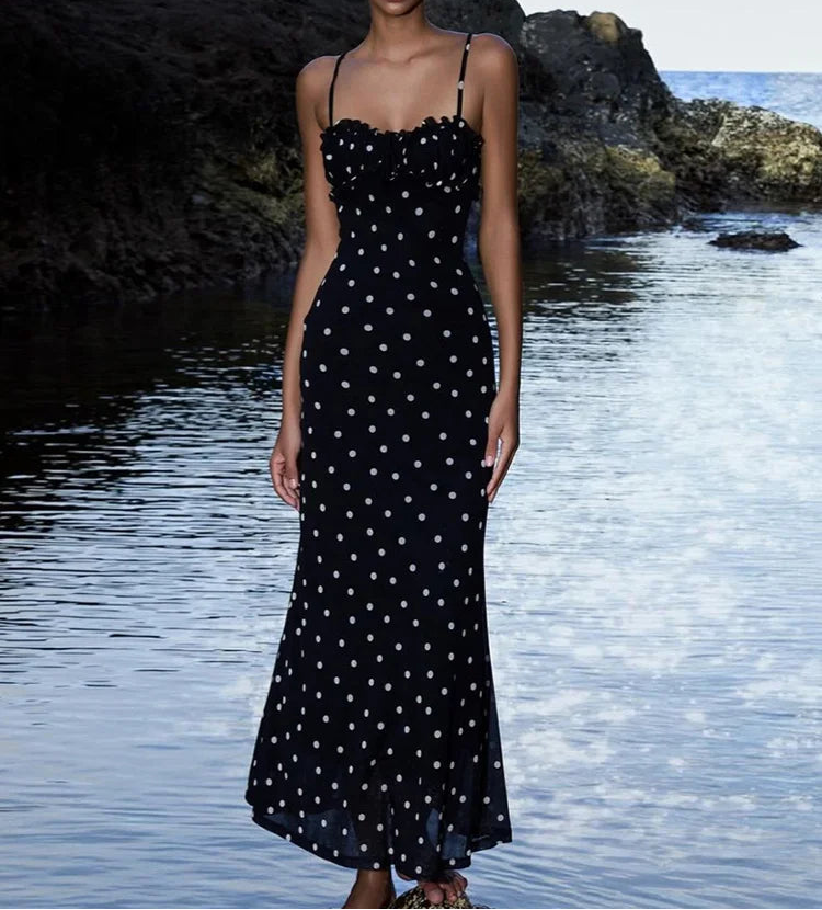 Woman in a black polka dot dress standing by a body of water with rocks.