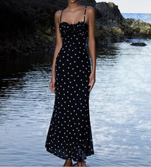Woman in a black polka dot dress standing by a body of water with rocks.