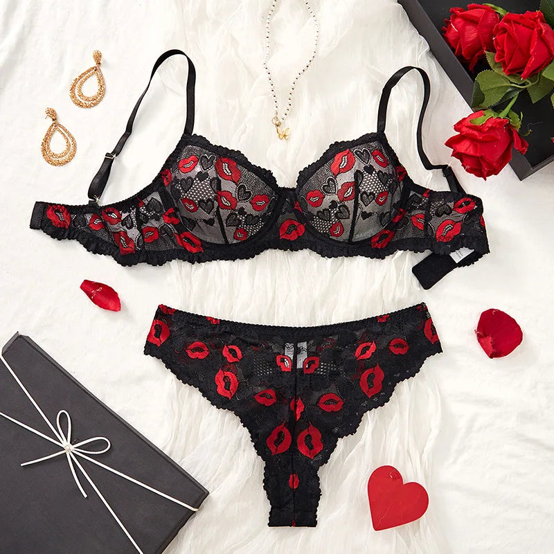 Black lace lingerie set with red accents on a white surface, surrounded by jewelry and flowers.