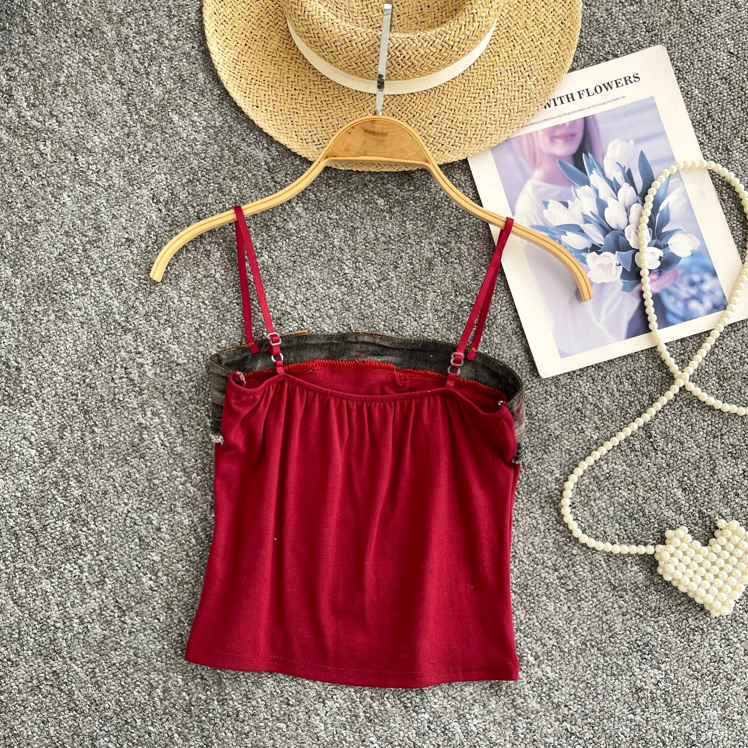 Red top on a hanger with a hat, newspaper, and pearl necklace on a gray surface