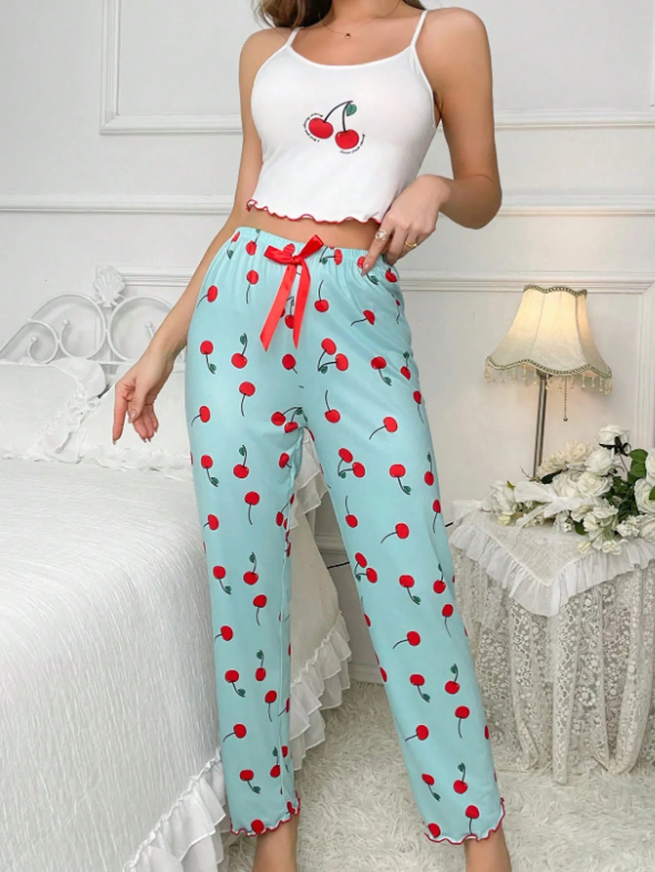 Woman wearing a white crop top with cherry design and light blue pants with red cherry pattern in a bedroom setting.