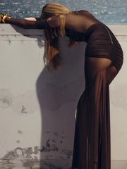 Woman in a brown dress leaning against a wall with water in the background