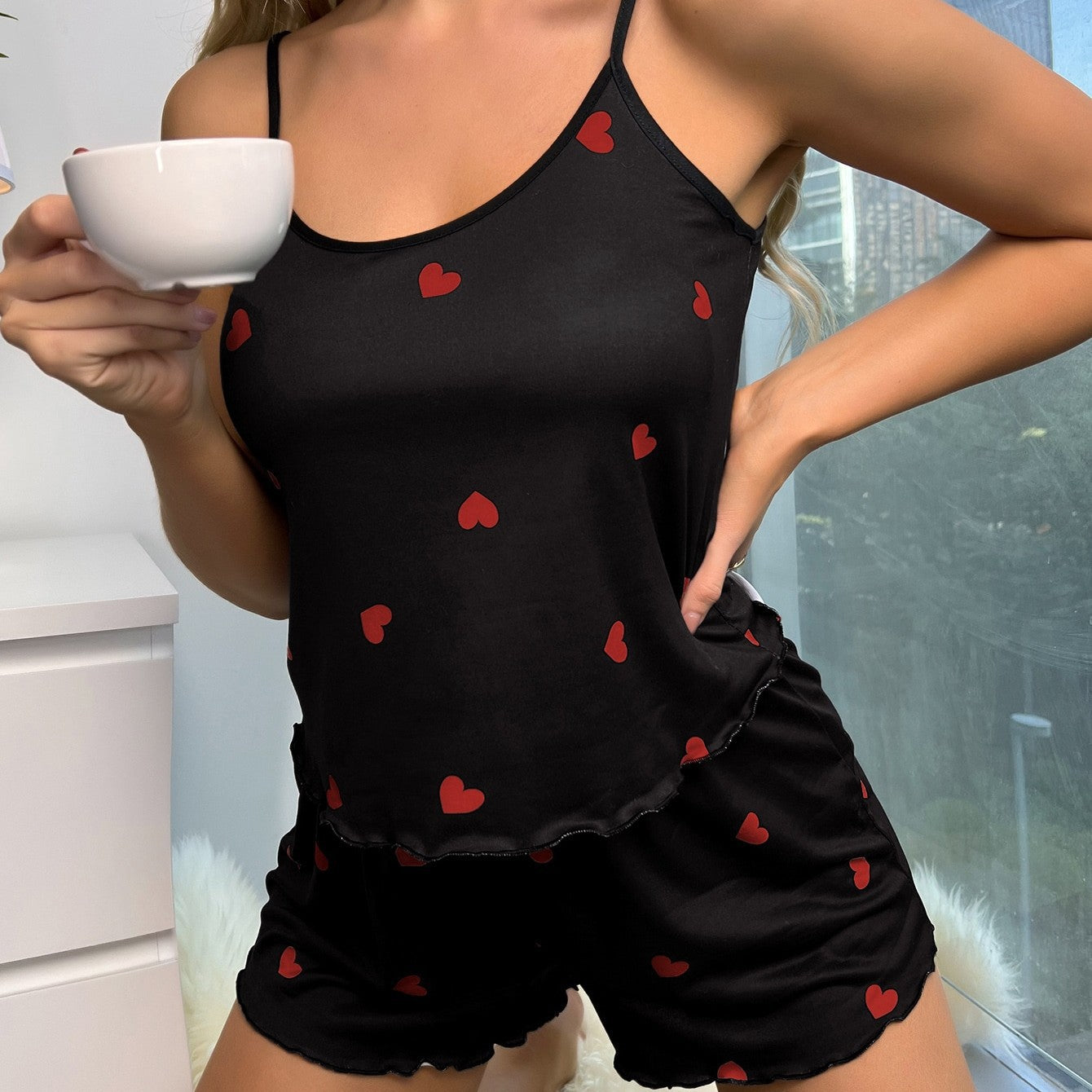 Person wearing a black tank top and shorts with red heart patterns, holding a white mug.