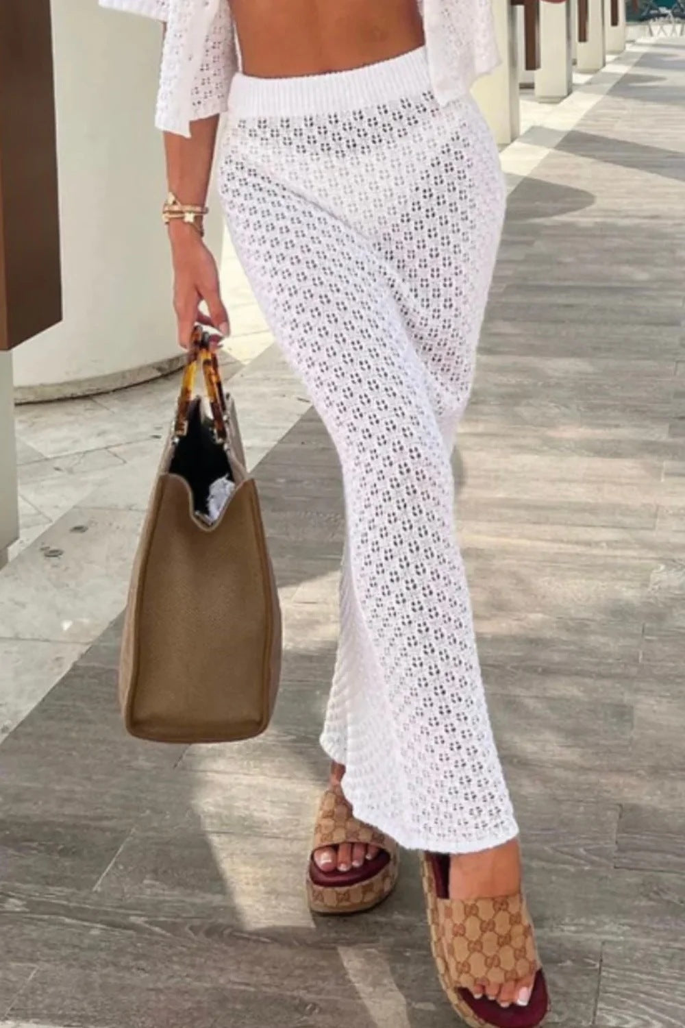 Person wearing a white lace skirt and sandals, holding a brown bag on a wooden floor.